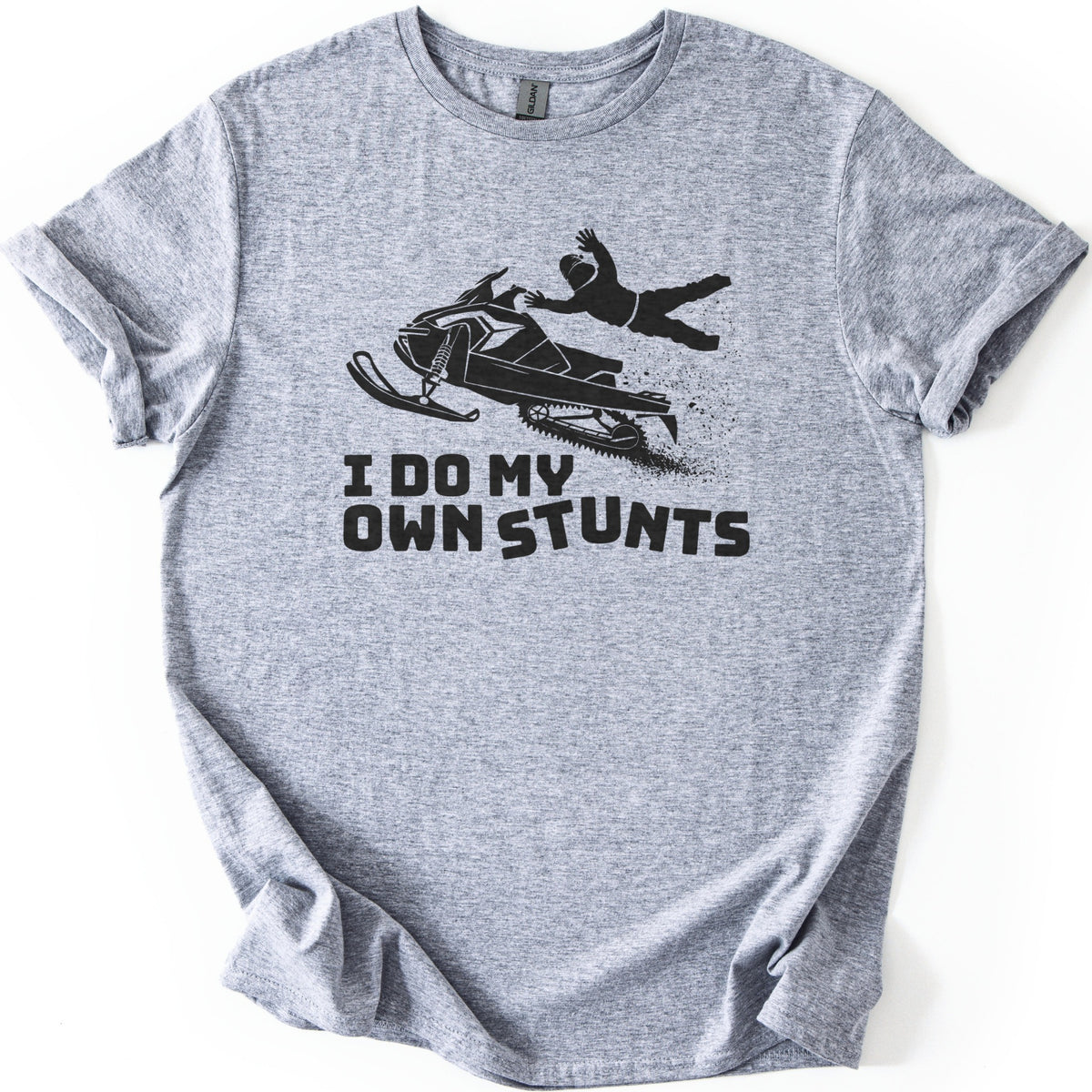 Snow Mobile Stunts Comfort Colors T-shirt in Sport Grey, laid flat. A black silhouette of a snowmobile flying through the air, its rider thrown off behind it in a spray of snow, above the words I Do My Own Stunts. The shirt lies on a white background with rolled sleeves.