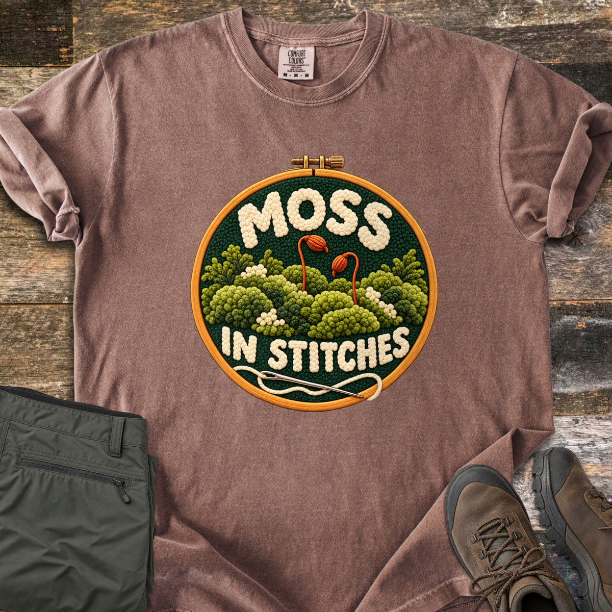 Moss in Stitches Comfort Colors T-shirt in Espresso, laid flat. An embroidery hoop holds stitched green moss with two orange spore capsules, the word Moss above and In Stitches below, plus a needle and thread. Folded olive pants and brown hiking boots lie beside it on weathered wood.