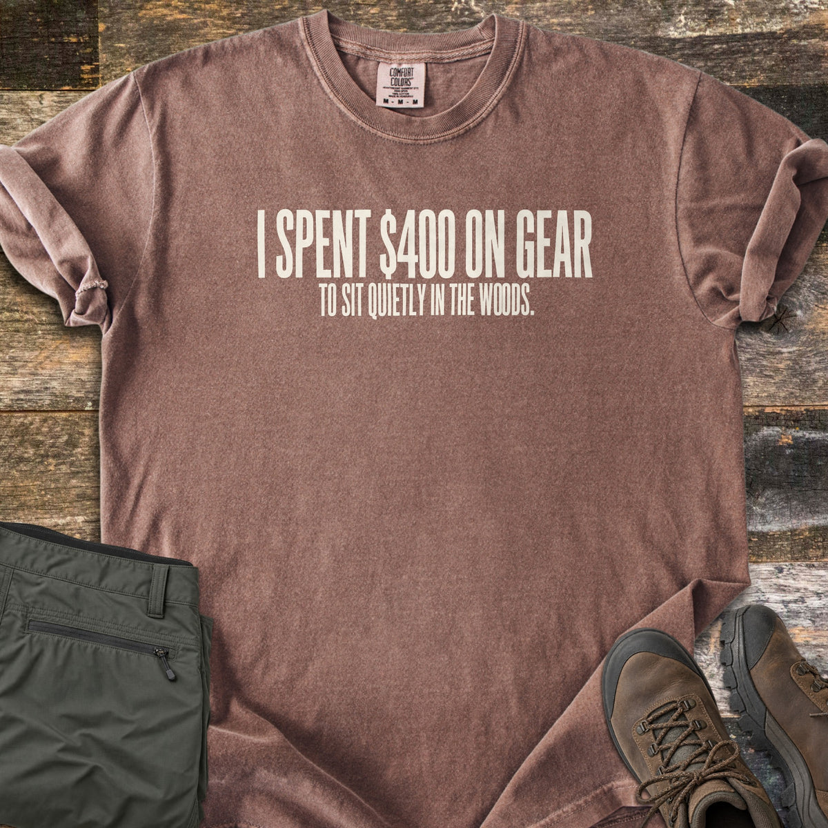 Gear To Sit Quietly Comfort Colors T-shirt in Espresso, laid flat. Cream text reads I SPENT $400 ON GEAR, with TO SIT QUIETLY IN THE WOODS. below. Sleeves are rolled on a rustic wood surface, beside folded olive hiking pants and brown hiking boots.