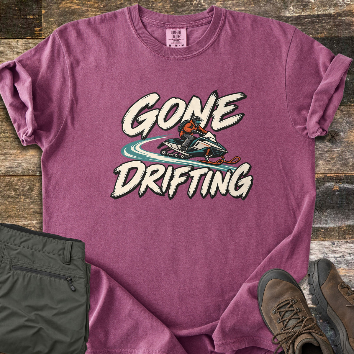 Gone Drifting Comfort Colors T-shirt in Berry, laid flat. Cream brush letters read Gone Drifting around a helmeted rider in an orange jacket on a teal snowmobile with a swirling snow trail. Sleeves rolled, on weathered wood with folded olive pants and brown hiking boots.