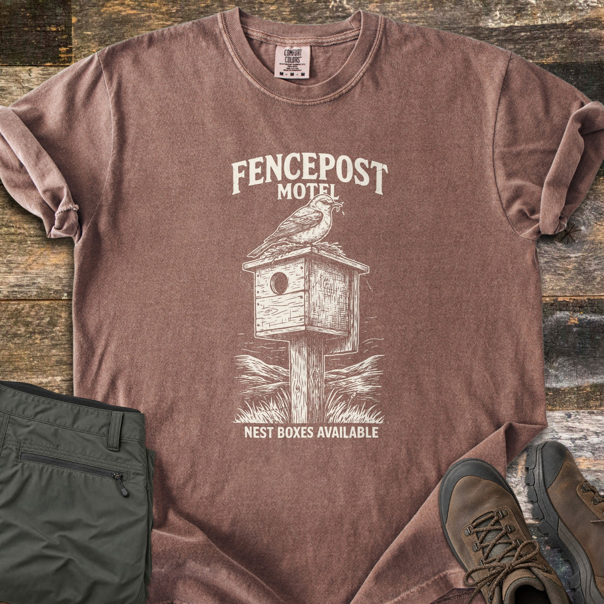 Fencepost Motel Comfort Colors T-shirt in Espresso, laid flat. A cream sketch of a bird perched on a wooden nest box atop a post in tall grass, with the words Fencepost Motel above and Nest Boxes Available below. Sleeves are rolled on a wood plank surface beside olive pants and brown hiking boots.