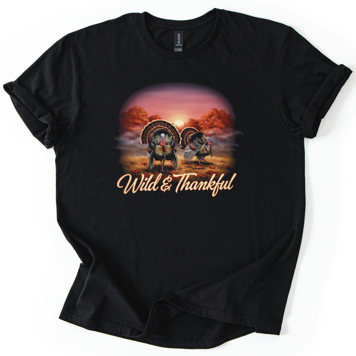 Wild & Thankful Comfort Colors T-shirt in Black, laid flat. Two wild turkeys with fanned tails stand in a grassy field under orange autumn trees at sunset, above the script words Wild & Thankful. The sleeves are rolled, on a white background.