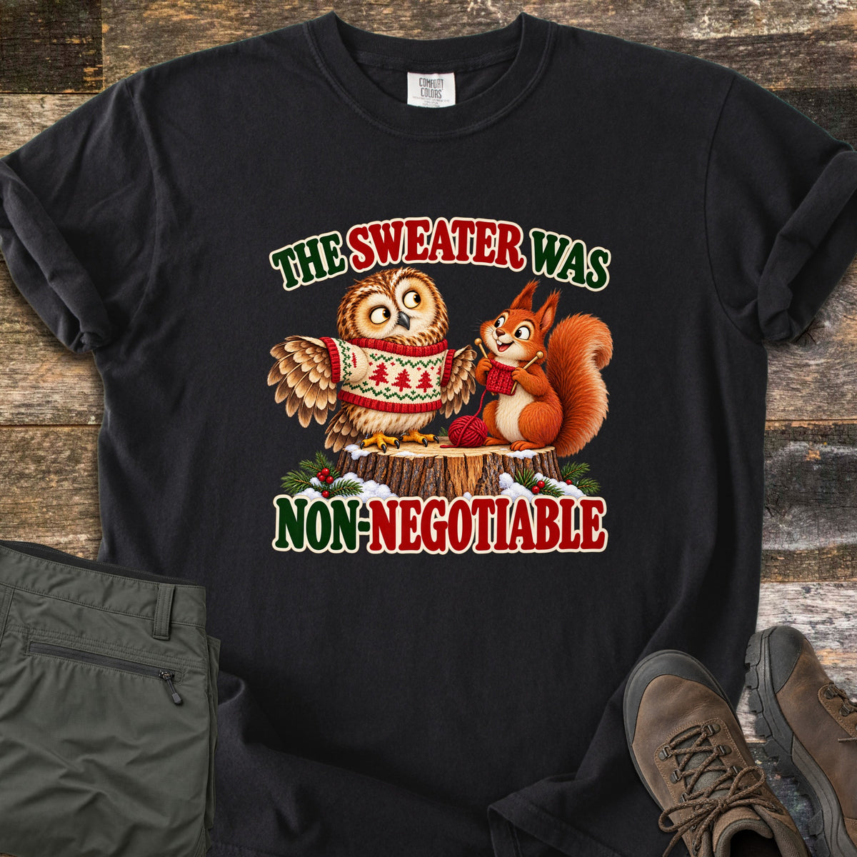 The Sweater Was Non-Negotiable Comfort Colors T-shirt in Black, laid flat. A cartoon owl in a red and cream Christmas tree sweater sits on a snowy stump beside a squirrel knitting with red yarn, between the words The Sweater Was Non-Negotiable. Olive pants and brown boots lie on weathered wood.