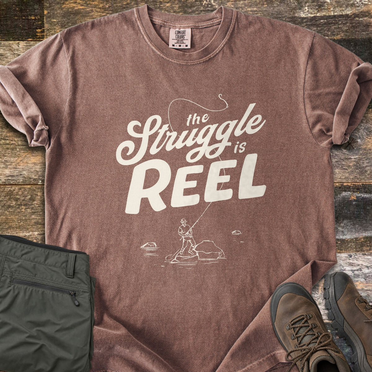 The Struggle Is Reel Comfort Colors T-shirt in Espresso, laid flat. Cream text reads the Struggle is REEL under an arcing fishing line with a hook, above a sketch of an angler casting from a rock in water. Folded olive pants and brown hiking boots sit on wood planks.