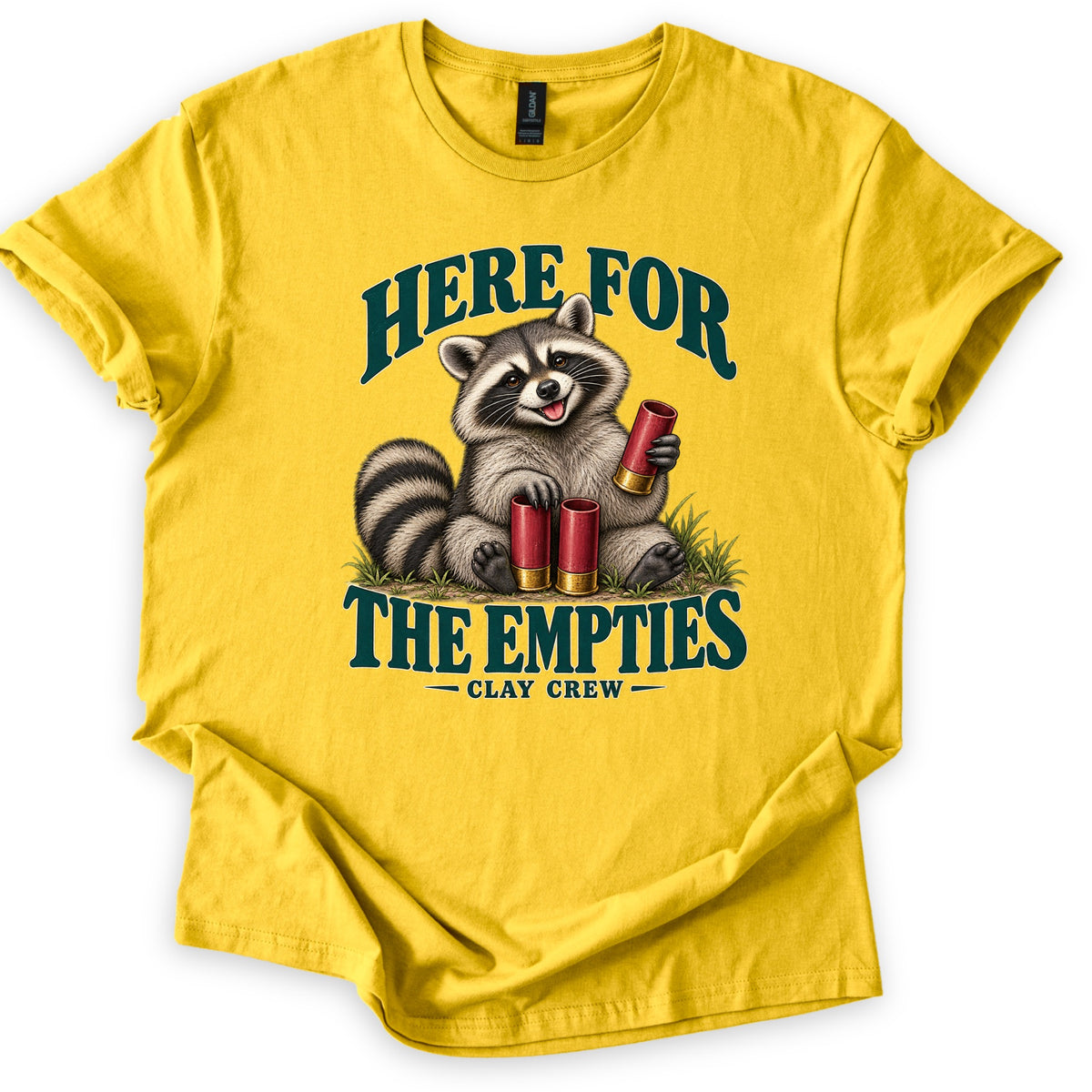 Here for the Empties Comfort Colors T-shirt in Daisy, laid flat. A grinning raccoon sits in grass holding a red shotgun shell, with two more at its feet, between the words Here For The Empties and Clay Crew in dark green. Sleeves are rolled, on a white background.