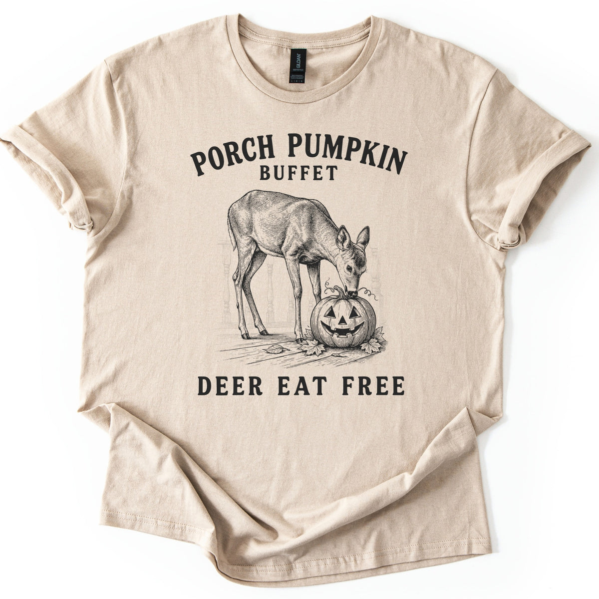 Porch Pumpkin Buffet Comfort Colors T-shirt in Sand, laid flat. A black engraving-style drawing of a deer nibbling a carved jack-o'-lantern on a porch with fallen leaves, between the words Porch Pumpkin Buffet and Deer Eat Free. Sleeves are rolled, on a white background.