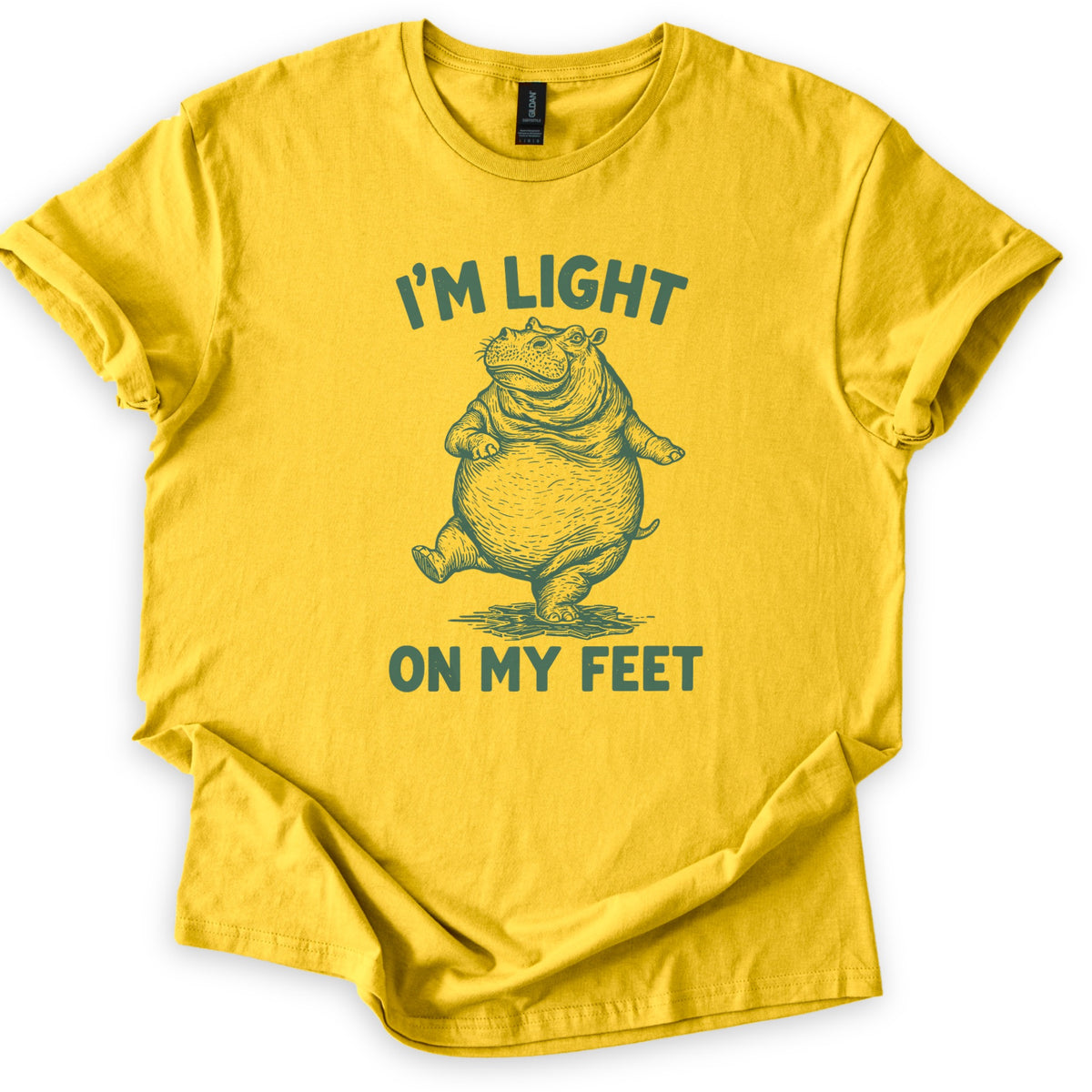 I'm Light On My Feet Comfort Colors T-shirt in Daisy, laid flat. A dark green etched-style drawing of a round hippo standing upright on one foot, mid-step, between the words I'M LIGHT above and ON MY FEET below. The shirt has rolled sleeves on a white background.