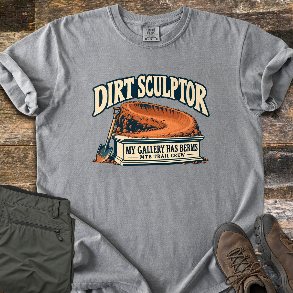 Dirt Sculptor Comfort Colors T-shirt in Grey, laid flat. The words Dirt Sculptor arch over a dirt berm on a stone pedestal reading My Gallery Has Berms, MTB Trail Crew, with a shovel leaning on it. Olive pants and brown hiking boots lie on wood planks.