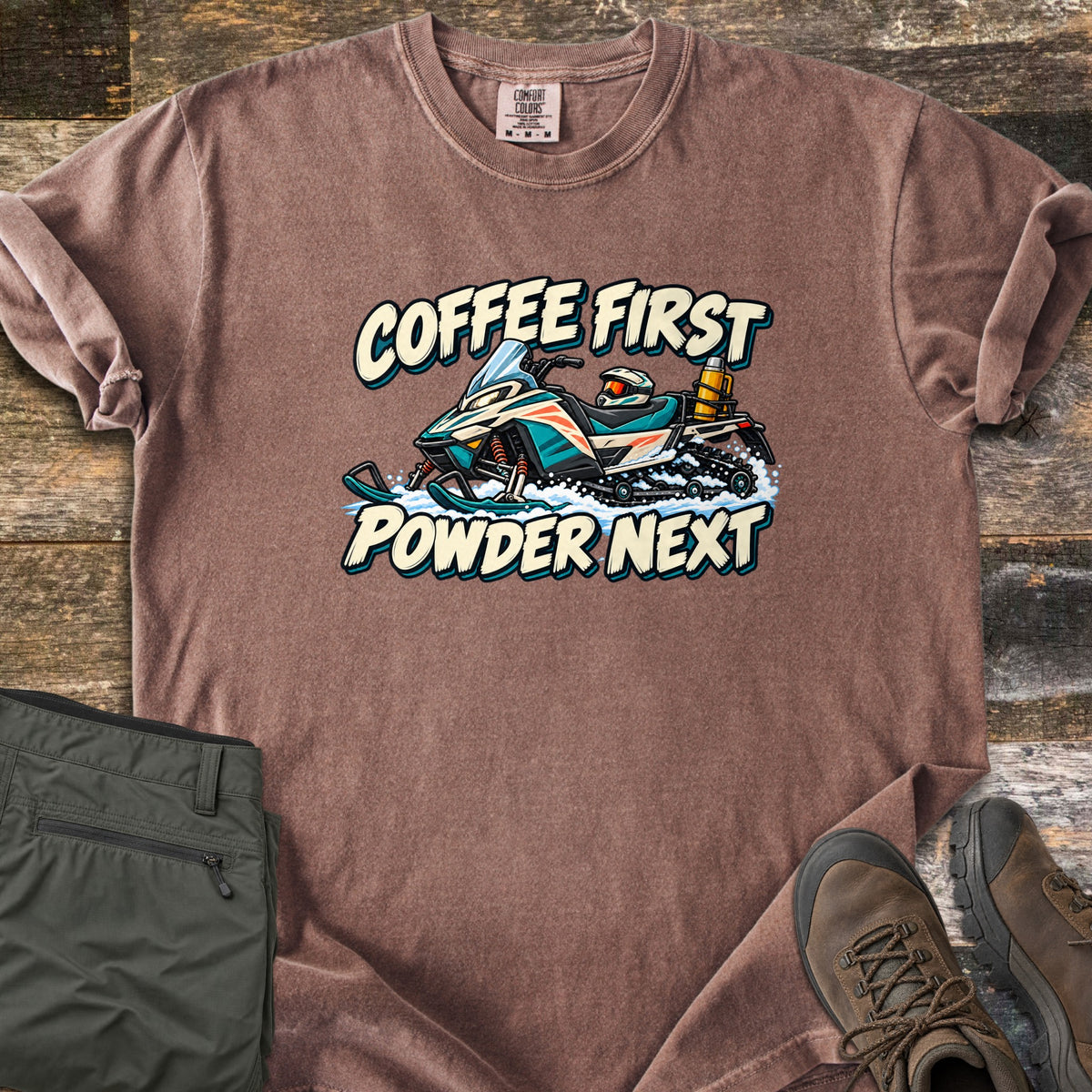 Coffee First Powder Next Comfort Colors T-shirt in Espresso, laid flat. A cartoon snowmobile with a helmet on the seat and a yellow thermos strapped on back kicks up snow between the words Coffee First Powder Next. Olive hiking pants and brown boots sit on a wood floor.