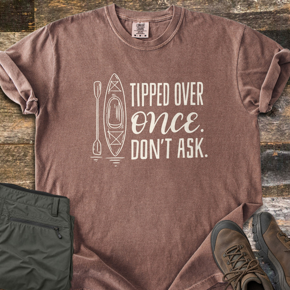 Tipped Over Once. Don't Ask. Comfort Colors T-shirt in Espresso, laid flat. A white line drawing of a kayak and paddle sits beside the words Tipped Over Once. Don't Ask., with Once in script. Sleeves rolled, on weathered wood with folded olive hiking pants and brown hiking boots.