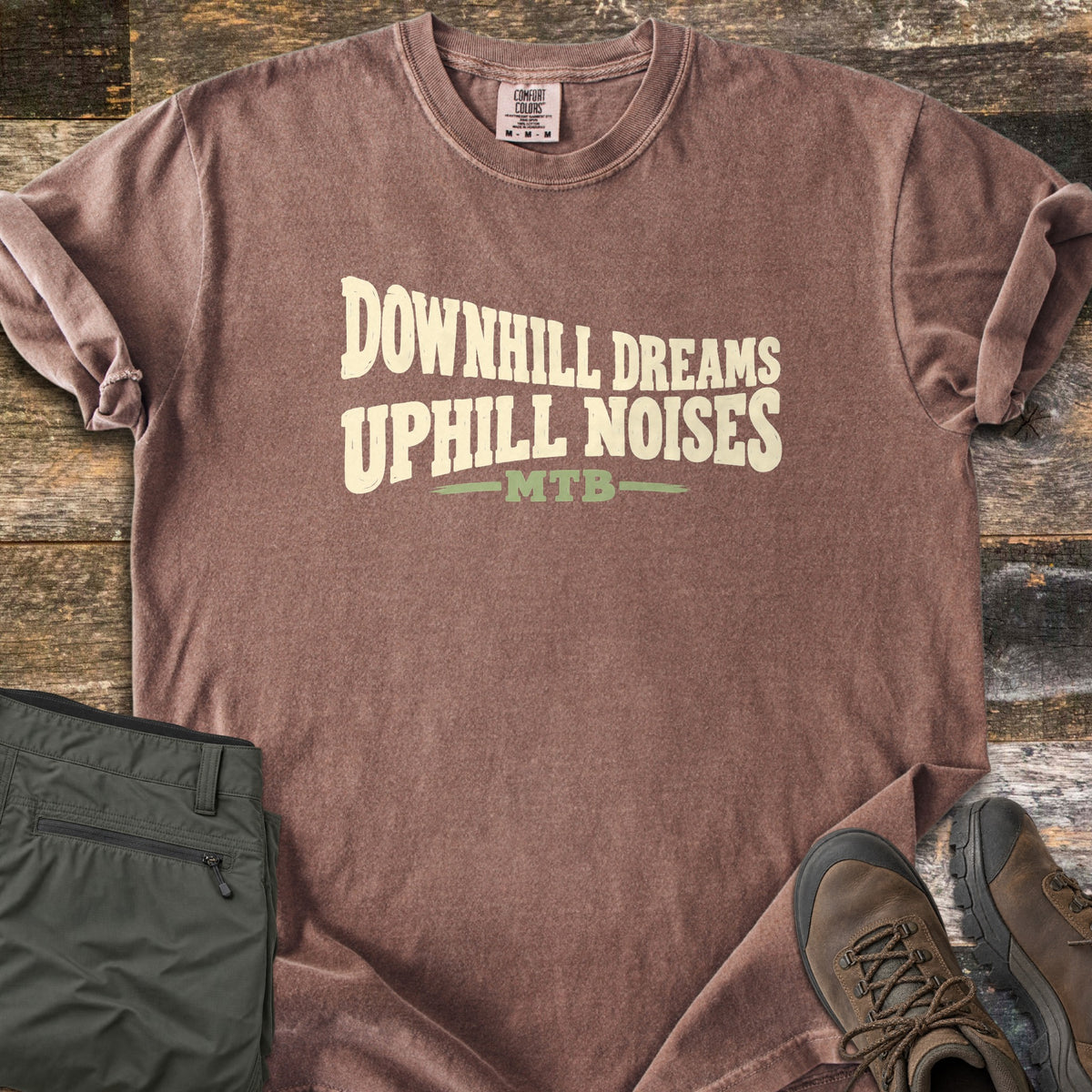 Downhill Dreams Uphill Noises Comfort Colors T-shirt in Espresso, laid flat. Cream wavy block letters read Downhill Dreams Uphill Noises above MTB in green between two green lines. Sleeves rolled on weathered wood planks, beside folded olive hiking pants and brown hiking boots.