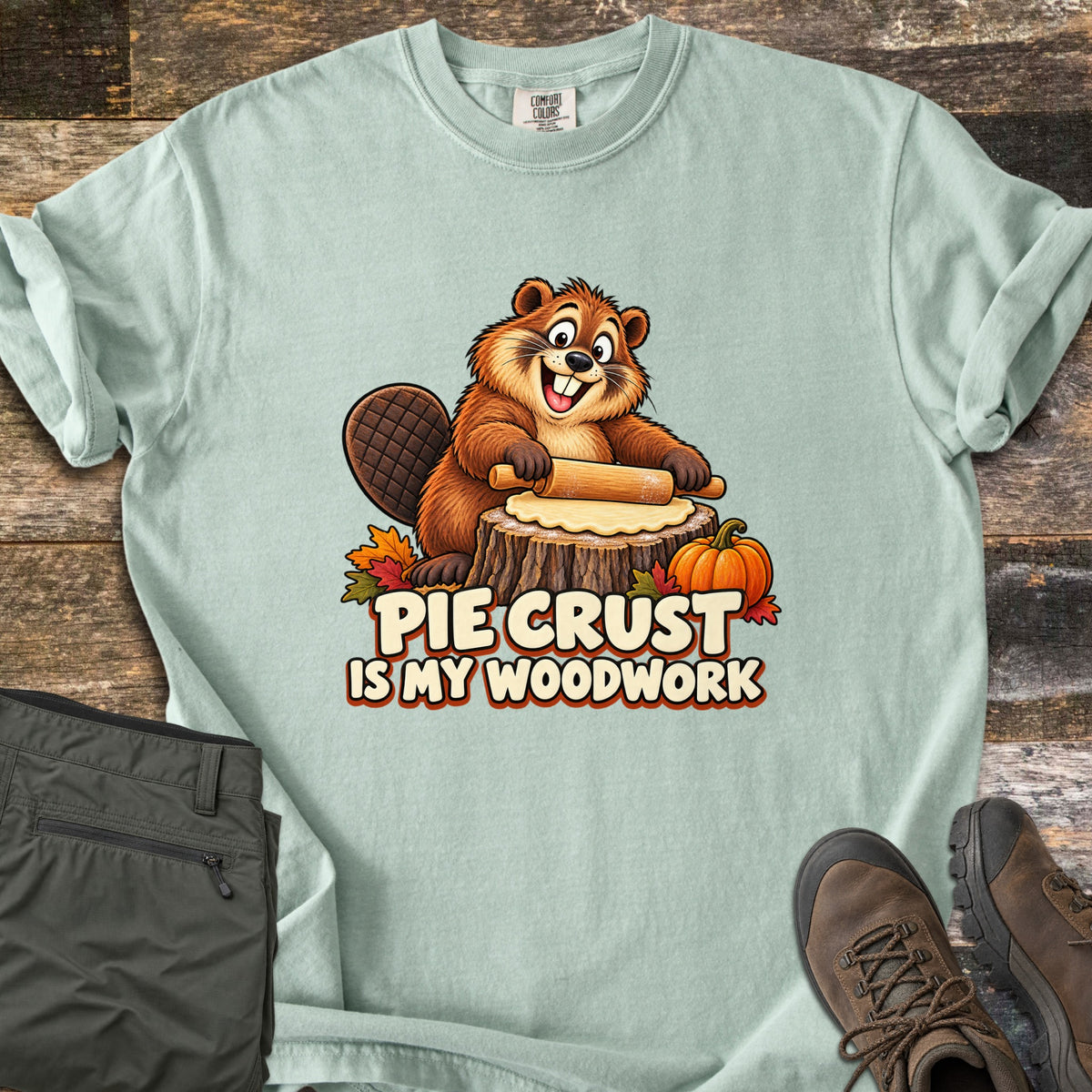 Pie Crust Is My Woodwork Comfort Colors T-shirt in Bay, laid flat. A grinning cartoon beaver rolls pie dough on a tree stump, with a small pumpkin and autumn leaves, above the words Pie Crust Is My Woodwork. Olive pants and brown hiking boots sit on a rustic wood surface.