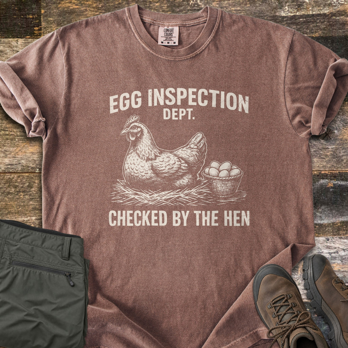 Egg Inspection Dept Comfort Colors T-shirt in Espresso, laid flat. A sketched hen sits on straw beside a basket of eggs, with Egg Inspection Dept. above and Checked By The Hen below in cream. Olive cargo pants and brown hiking boots lie on a rustic wood floor.
