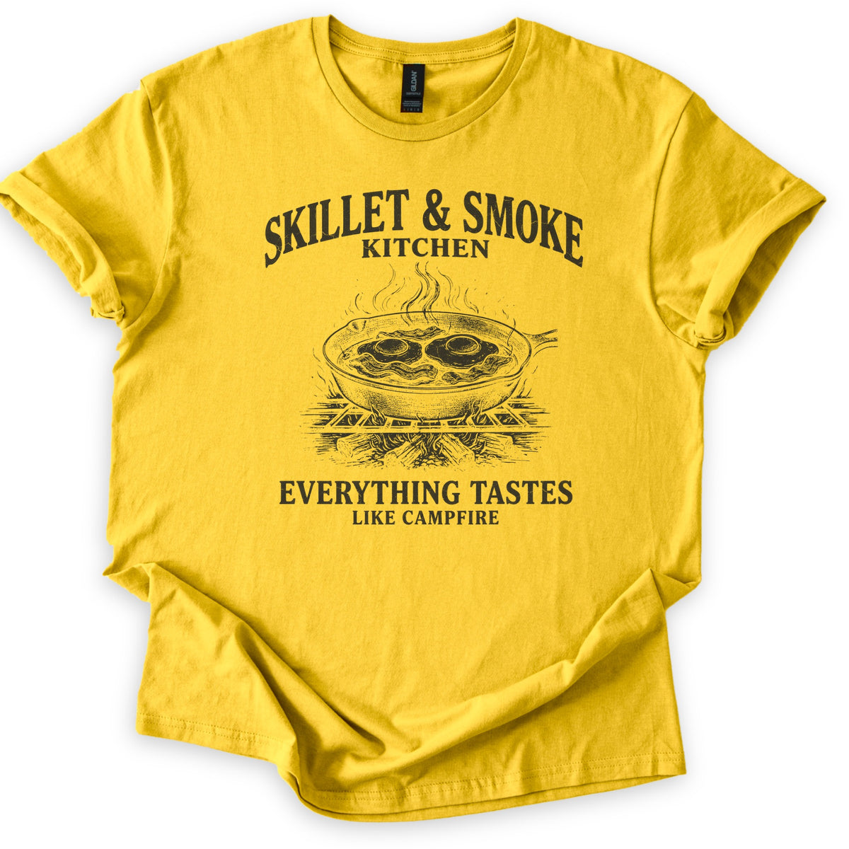 Skillet & Smoke Kitchen Comfort Colors T-shirt in Daisy, laid flat. A black print shows a cast iron skillet of fried eggs and bacon steaming on a grate over a campfire, under the words Skillet & Smoke Kitchen and above Everything Tastes Like Campfire. Sleeves cuffed, on white.