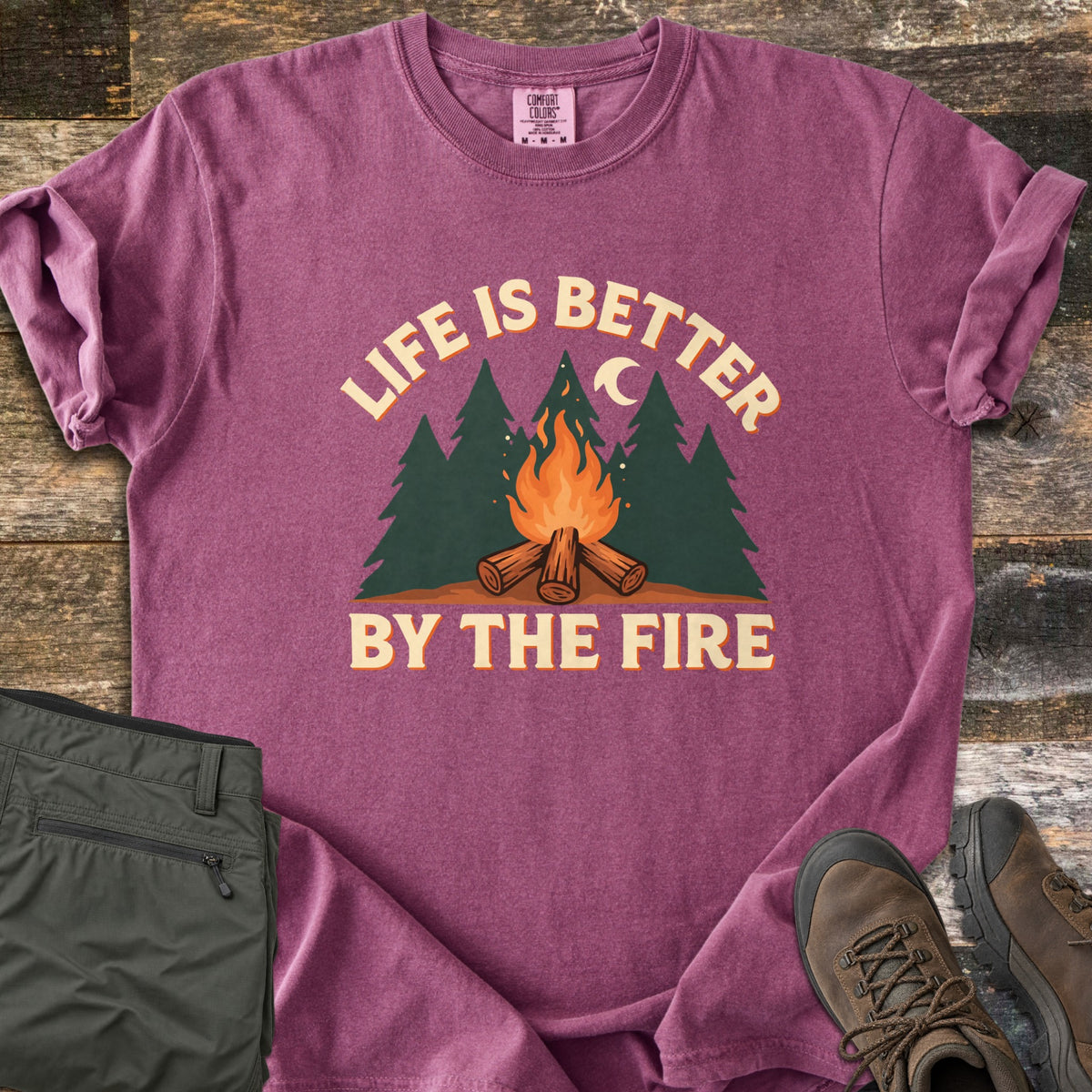Life Is Better By The Fire Comfort Colors T-shirt in Berry, laid flat. Arched cream text reads Life Is Better over dark green pine trees, a crescent moon and a campfire on crossed logs, with By The Fire below. Folded olive pants and brown hiking boots sit on weathered wood.