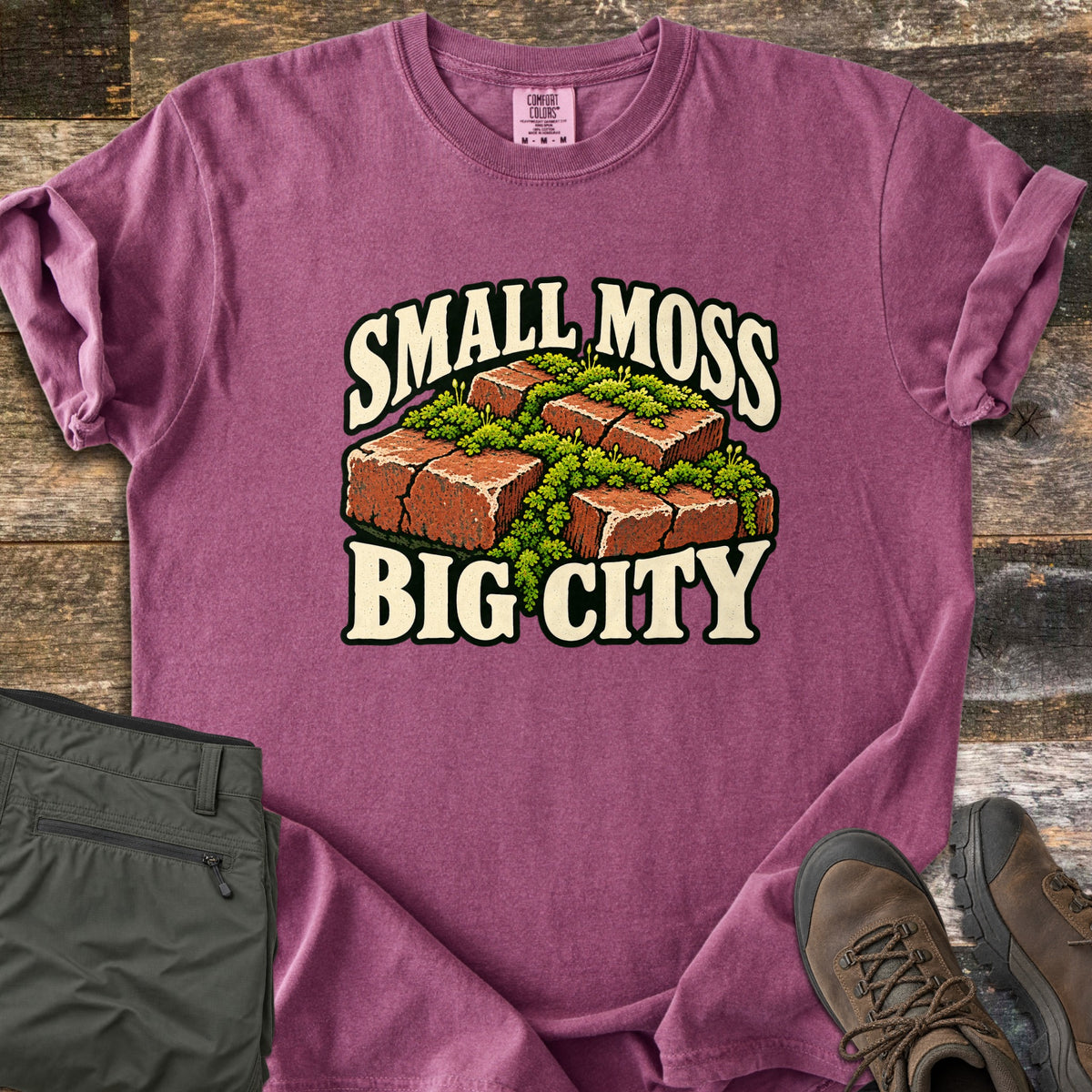 Small Moss Big City Comfort Colors T-shirt in Berry, laid flat. Red bricks with bright green moss growing between and over them, with the words Small Moss Big City in cream lettering. Sleeves are rolled, on a wood floor beside folded olive pants and brown hiking boots.