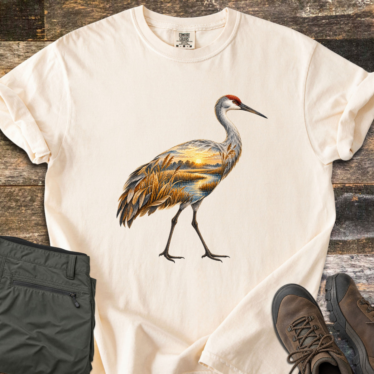 Crane Marsh Sunrise Comfort Colors T-shirt in Ivory, laid flat. A sandhill crane with a red crown walks across the chest, its body filled with a sunrise marsh scene of golden reeds and a winding stream. Rolled sleeves; gray pants and brown hiking boots sit on weathered wood.