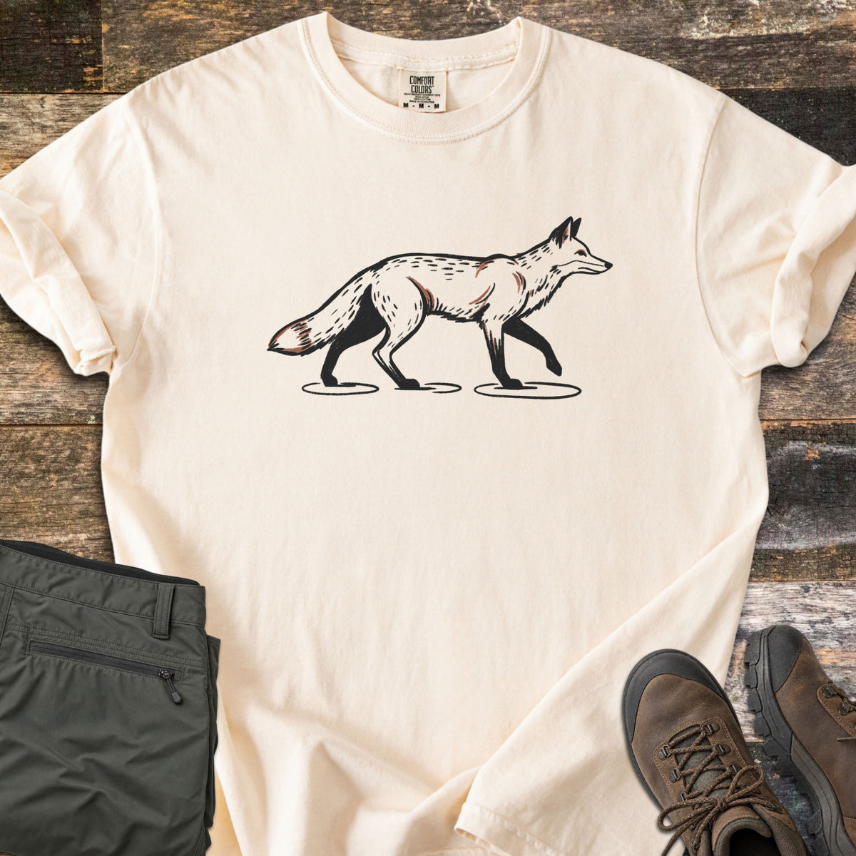 Trotting Fox Comfort Colors T-shirt in Ivory, laid flat. A black-outlined illustration of a pale fox with rust shading trotting to the right across small ground patches, printed on the chest with no words. The shirt lies on weathered wood beside olive green hiking pants and brown hiking boots.