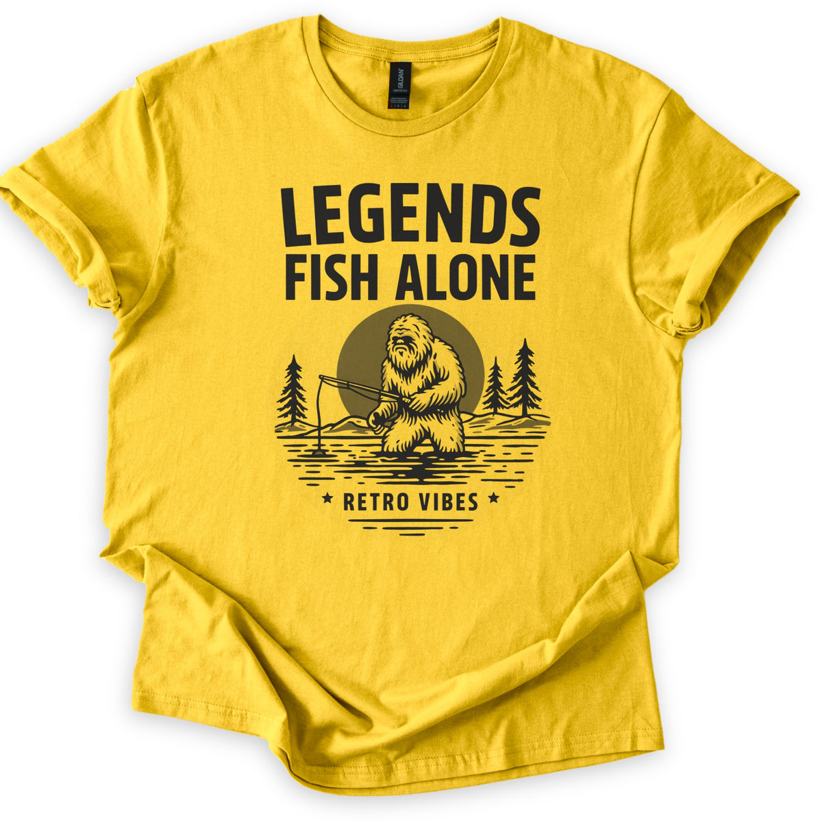 Legends Fish Alone Bigfoot Comfort Colors T-shirt in Daisy, laid flat. Black print of Bigfoot fishing knee-deep in a lake before an olive moon and pine trees, under the words Legends Fish Alone and above Retro Vibes between two stars. Sleeves cuffed, on a white background.