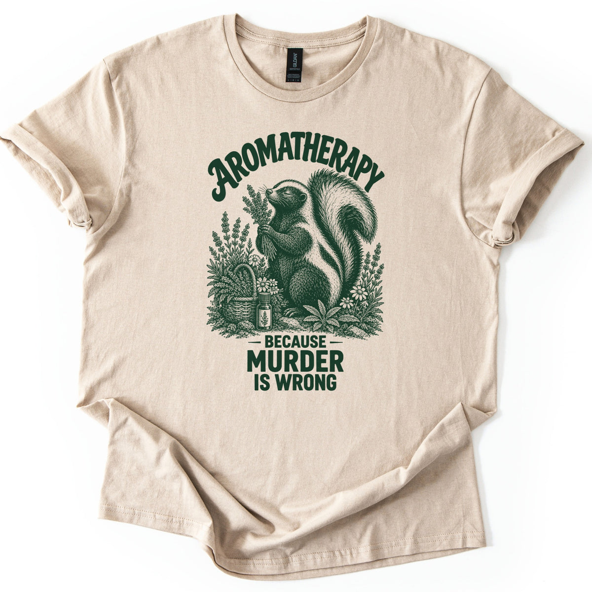 Aromatherapy Murder Skunk Comfort Colors T-shirt in Sand, laid flat. A dark green skunk sniffs a lavender bundle beside a basket, flowers and an oil bottle, under the word Aromatherapy and above Because Murder Is Wrong. Sleeves are cuffed on a white background.