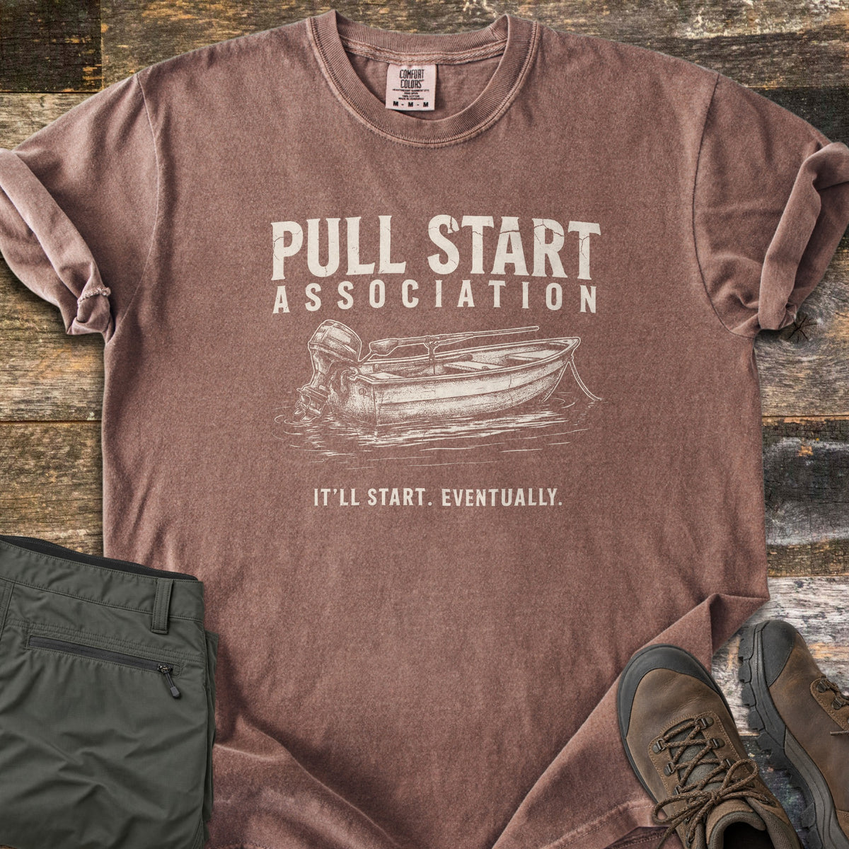 Pull Start Association Comfort Colors T-shirt in Espresso, laid flat. Cream distressed text reads Pull Start Association above a sketched rowboat with an outboard motor on water, and below it It'll Start. Eventually. Sleeves are rolled on a wood floor beside green pants and brown hiking boots.