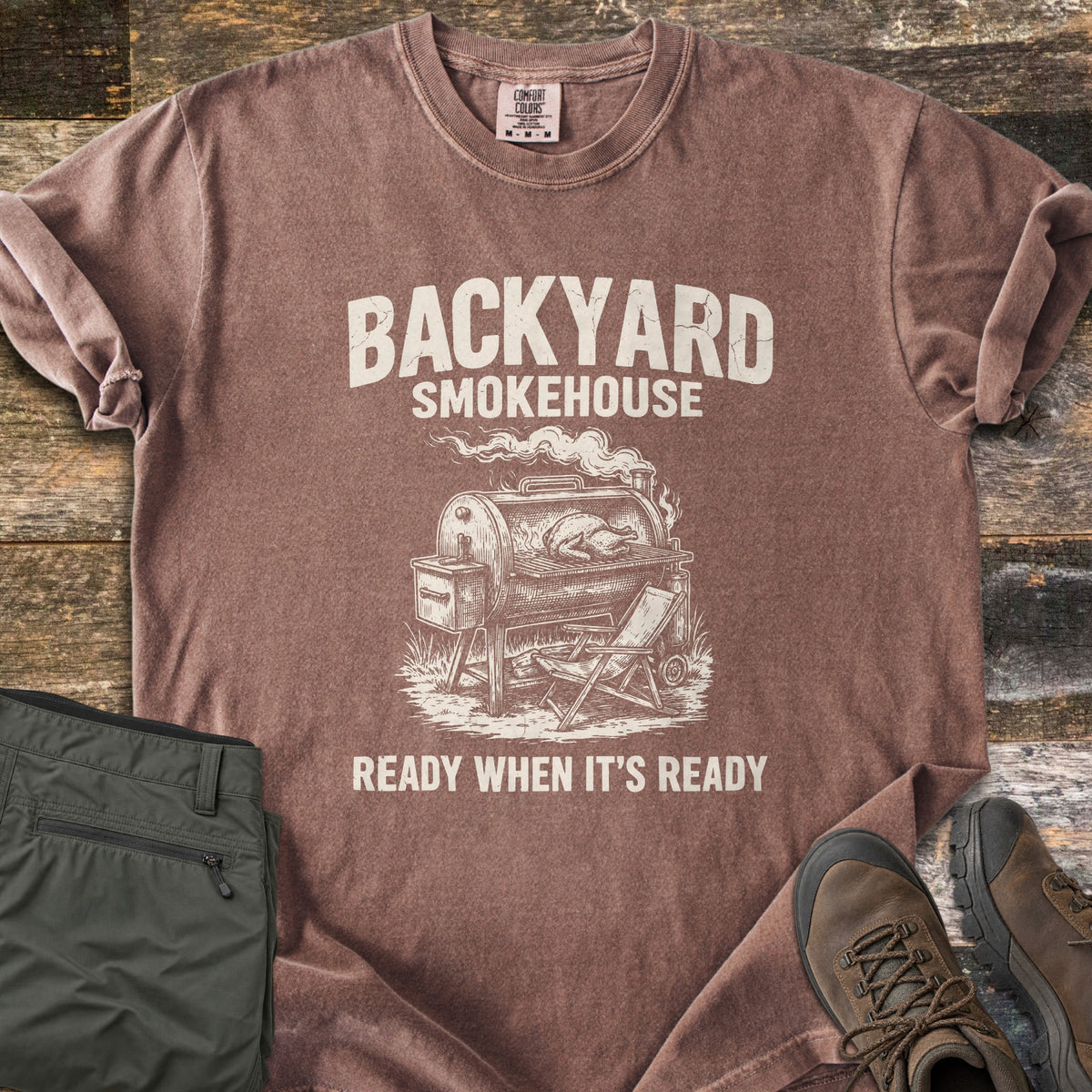 Backyard Smokehouse Comfort Colors T-shirt in Espresso, laid flat. Cream print of a smoker grill with a chicken inside and a folding chair beside it, under the words Backyard Smokehouse and above Ready When It's Ready. Olive pants and brown boots lie on weathered wood.