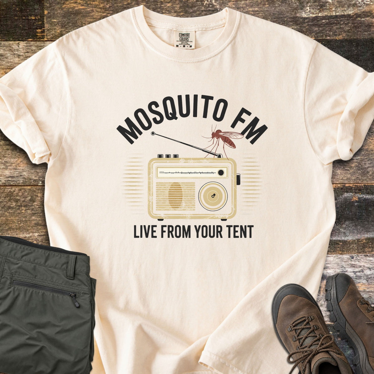 Mosquito FM Comfort Colors T-shirt in Ivory, laid flat. A mosquito perches on the antenna of a vintage tan radio under the arched black words Mosquito FM, with Live From Your Tent below. Set on weathered wood with folded olive hiking pants and brown hiking boots.