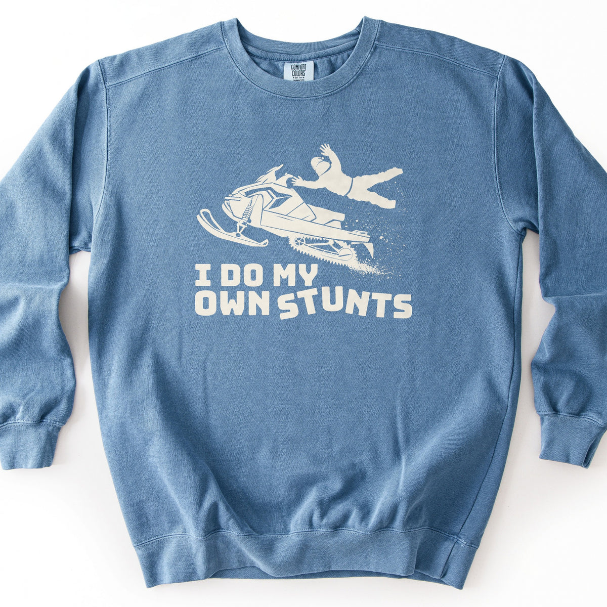 I Do My Own Stunts Comfort Colors crewneck sweatshirt in Blue Jean, laid flat. A cream print shows a helmeted rider thrown from a snowmobile in midair with a spray of snow, above the words I DO MY OWN STUNTS. The sweatshirt lies on a white surface.