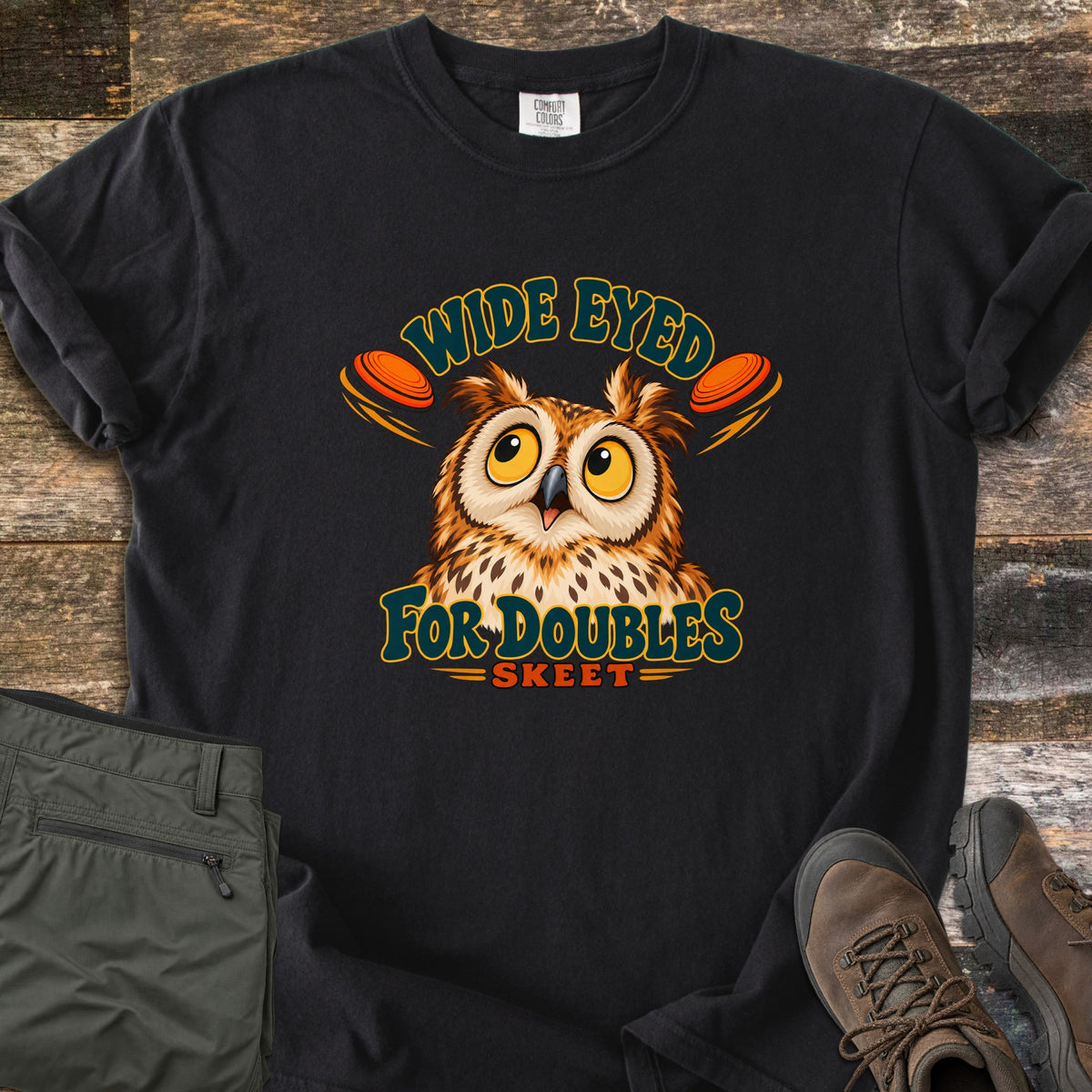 Wide Eyed for Doubles Comfort Colors T-shirt in Black, laid flat. A wide-eyed cartoon owl sits between two orange clay targets under the arched words Wide Eyed, with For Doubles and Skeet below. Sleeves are rolled, on weathered wood with olive pants and brown hiking boots.