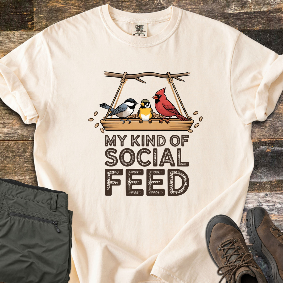My Kind Of Social Feed Comfort Colors T-shirt in Ivory, laid flat. A chickadee, a small yellow bird and a red cardinal sit on a rope-hung tray feeder full of seeds above the words My Kind Of Social Feed in brown. Olive hiking pants and brown hiking boots lie on weathered wood.