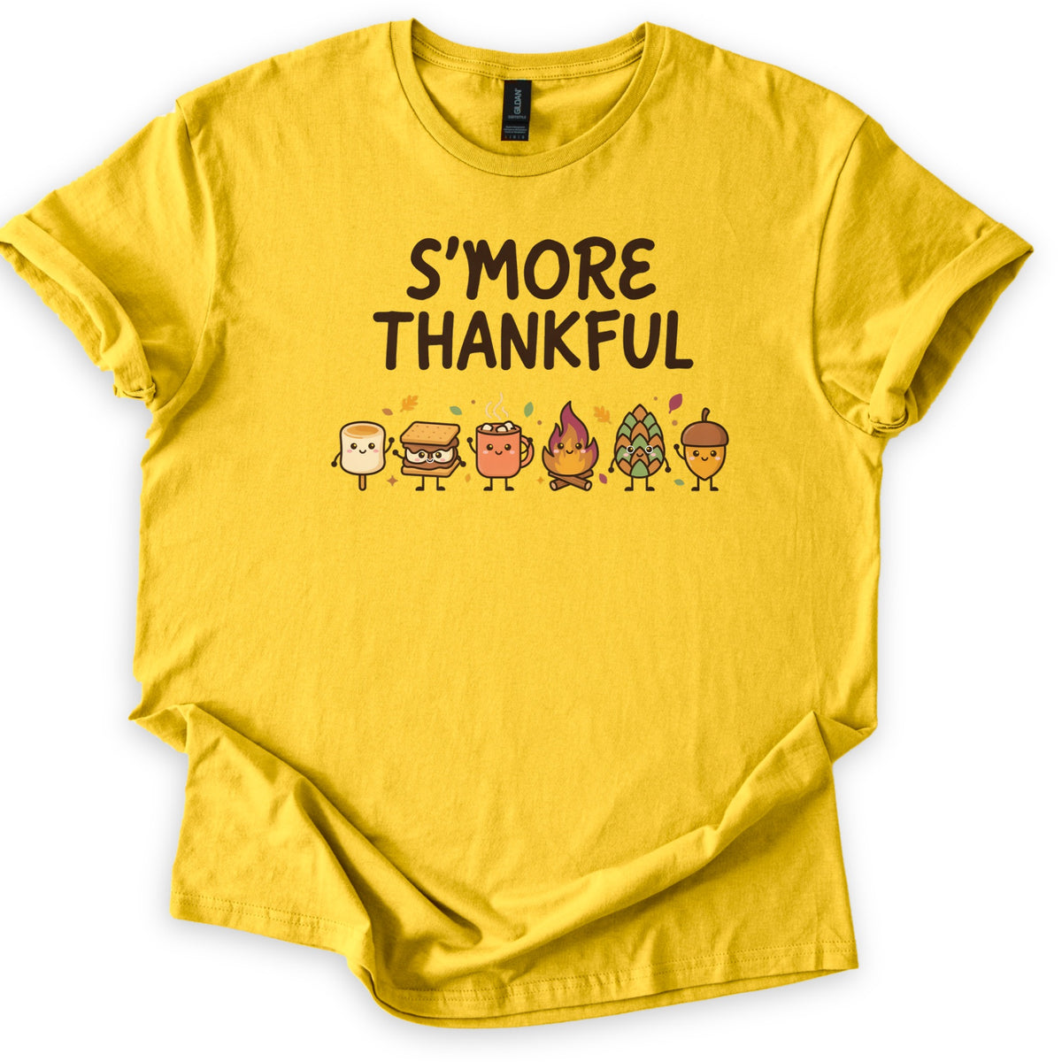 Smore Thankful Comfort Colors T-shirt in Daisy, laid flat. Dark brown letters read S'MORE THANKFUL above a row of smiling cartoon characters: a marshmallow, a s'more, a cocoa mug, a campfire, a pinecone and an acorn among autumn leaves. Sleeves rolled, on white.