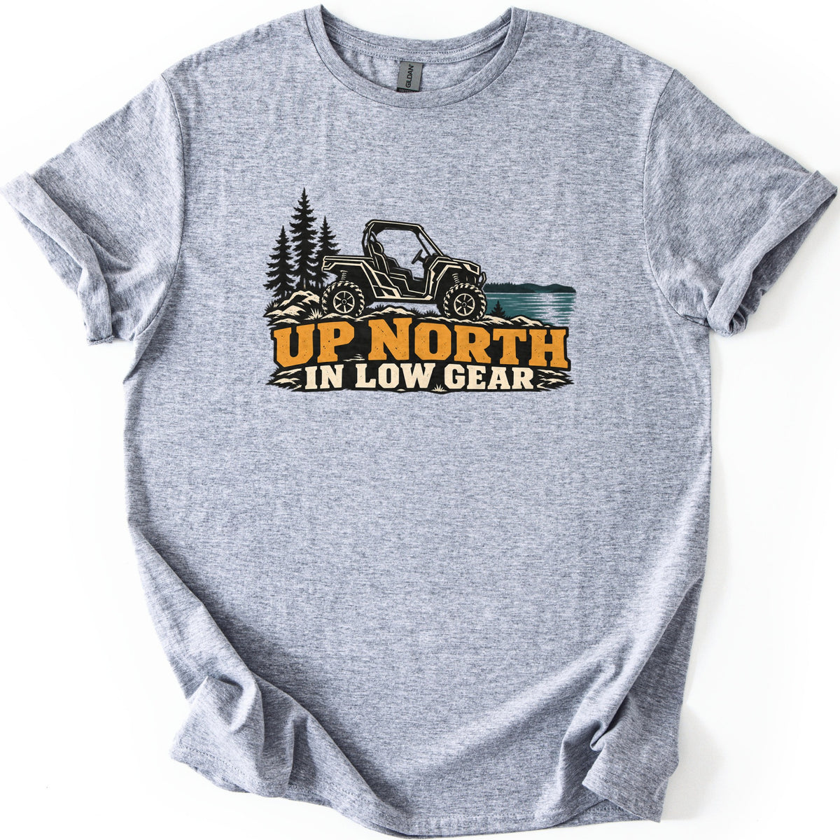 Up North in Low Gear Comfort Colors T-shirt in Sport Grey, laid flat. A black side-by-side UTV on rocky ground between pine trees and a blue lake sits above the words UP NORTH in orange and IN LOW GEAR in cream. The shirt has rolled sleeves on a white background.