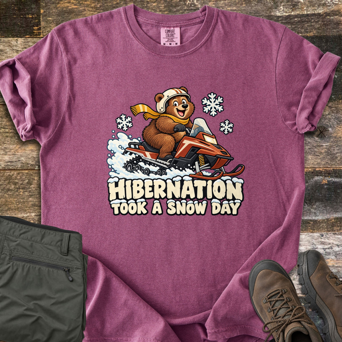 Hibernation Took a Snow Day Comfort Colors T-shirt in Berry, laid flat. A cartoon bear in a white helmet and yellow scarf rides a red snowmobile past snowflakes above the words Hibernation Took a Snow Day. Olive pants and brown hiking boots lie on rustic wood.