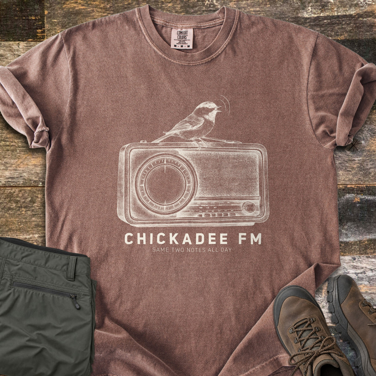 Chickadee FM Comfort Colors T-shirt in Espresso, laid flat. A sketched chickadee singing atop a vintage radio, above the words CHICKADEE FM and Same Two Notes All Day in cream. Sleeves rolled, on weathered wood with folded green hiking pants and brown hiking boots.