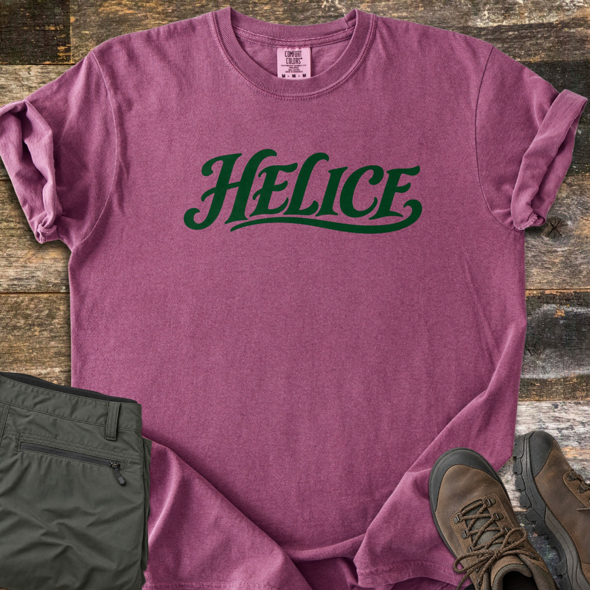Helice Comfort Colors T-shirt in Berry, laid flat. The word Helice is printed in dark green script lettering with a swash underline across the chest. Sleeves are rolled, on a rustic wood floor beside folded olive cargo pants and brown leather hiking boots.