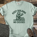 Keep Building Beaver Comfort Colors T-shirt in Bay, laid flat. A sketch-style beaver hugs a log beside a stack of logs, printed in dark green with the words Keep Building above and No Excuses below. Olive cargo pants and brown hiking boots lie beside it on weathered wood.