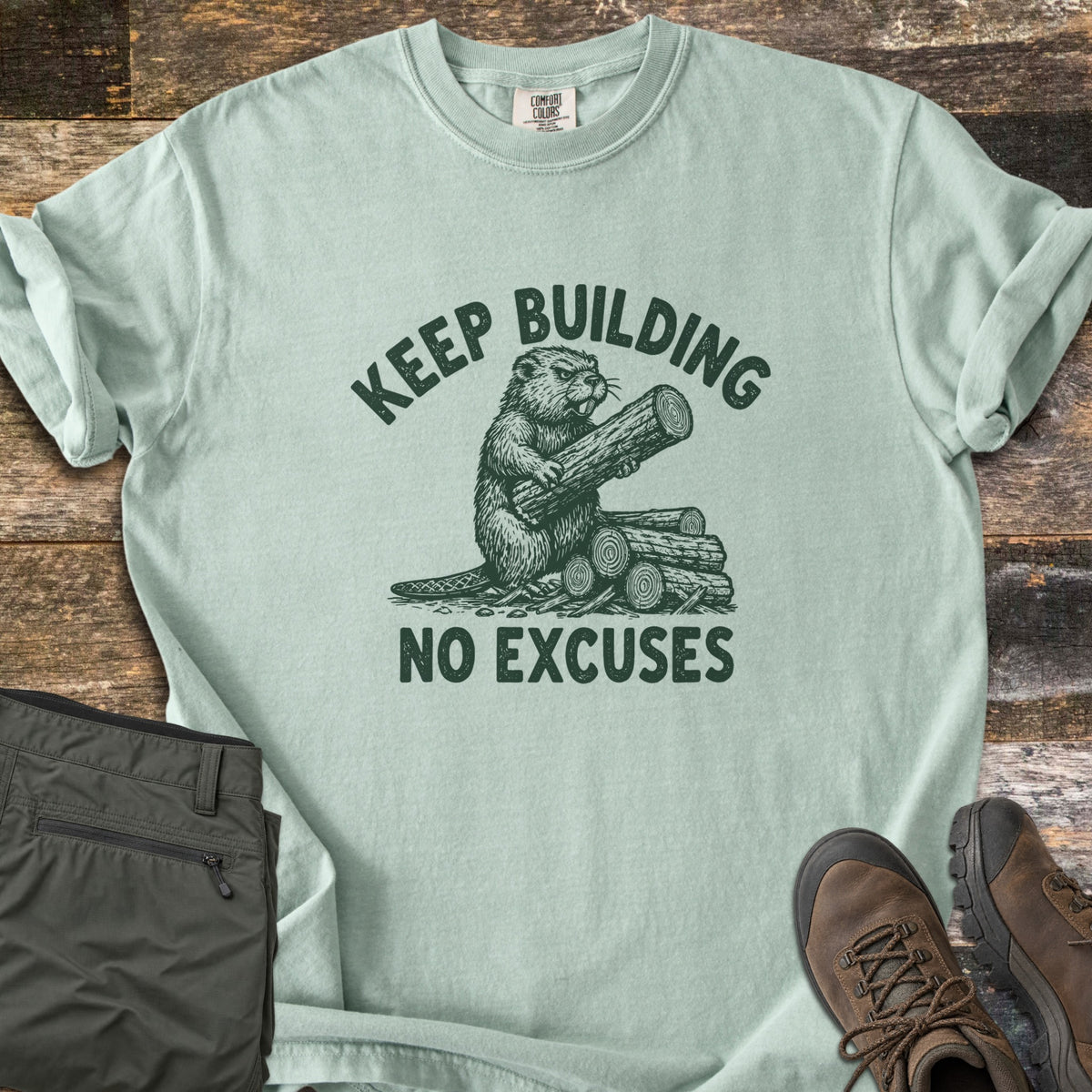 Keep Building Beaver Comfort Colors T-shirt in Bay, laid flat. A sketch-style beaver hugs a log beside a stack of logs, printed in dark green with the words Keep Building above and No Excuses below. Olive cargo pants and brown hiking boots lie beside it on weathered wood.