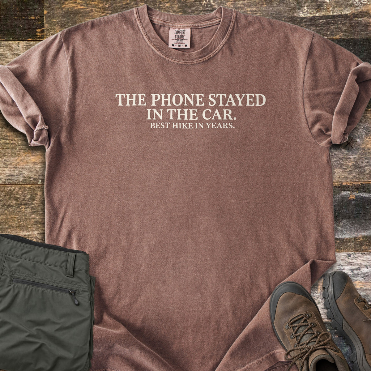 Phone Stayed In The Car Comfort Colors T-shirt in Espresso, laid flat. Cream serif text reads THE PHONE STAYED IN THE CAR. with BEST HIKE IN YEARS. in smaller letters below. Sleeves are rolled on a wood plank surface beside olive hiking pants and brown hiking boots.