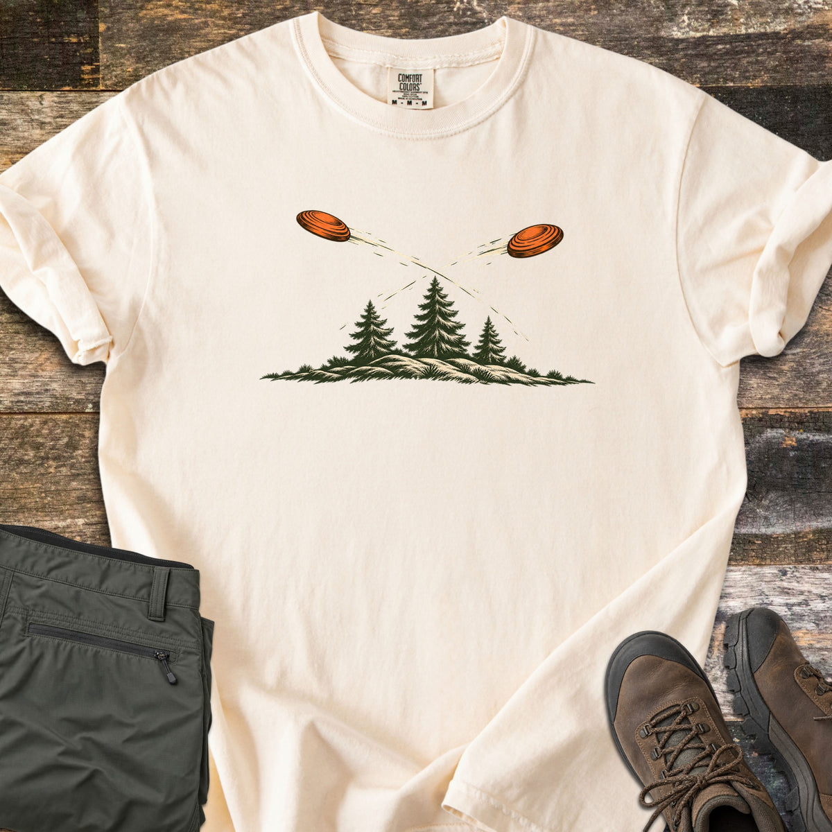 Crossing Clays Comfort Colors T-shirt in Ivory, laid flat. Two orange clay targets fly across each other, trailing streaks, above three pine trees on a grassy hill, with no words. The shirt lies on weathered wood beside folded olive pants and brown hiking boots.