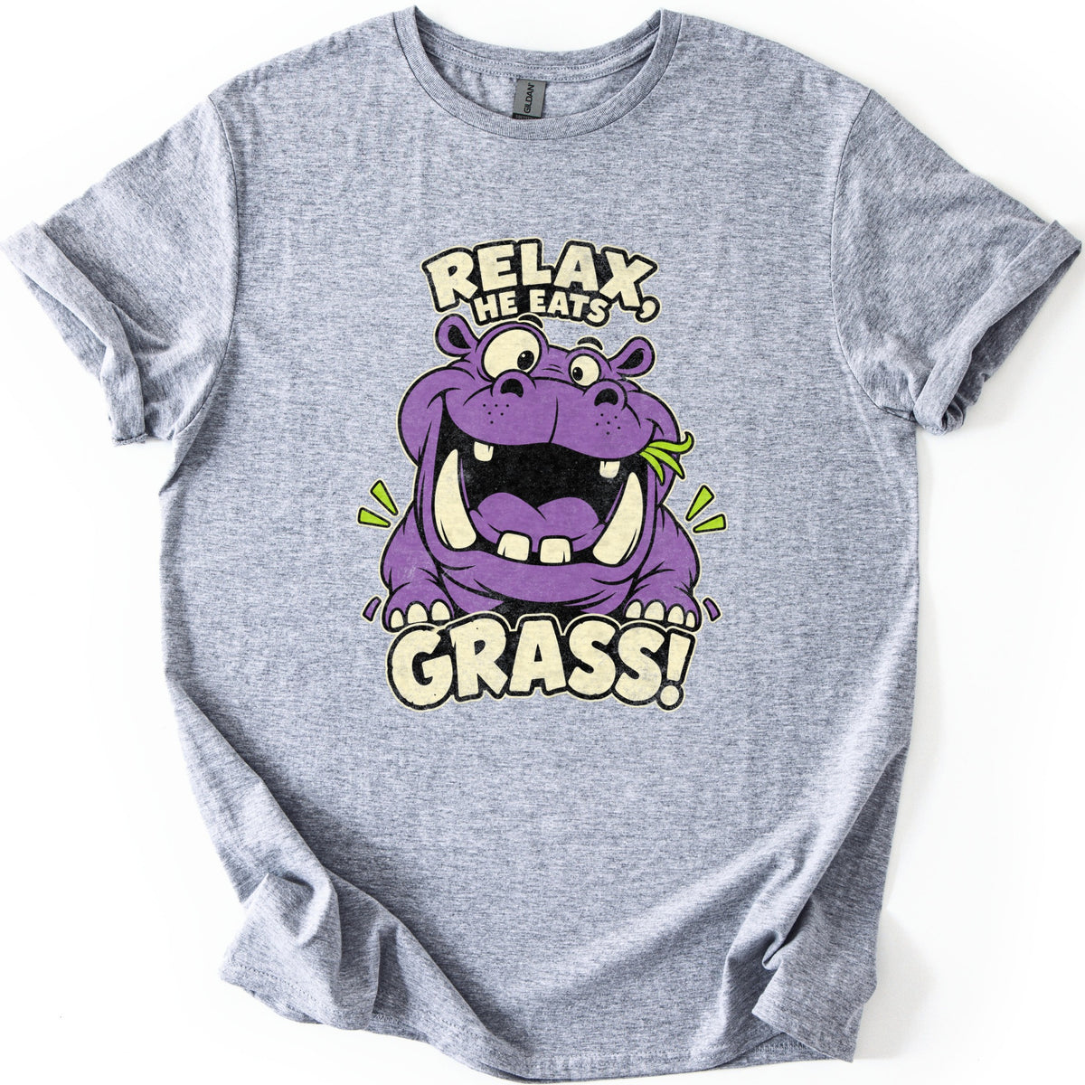 Relax He Eats Grass Comfort Colors T-shirt in Sport Grey, laid flat. A grinning purple cartoon hippo with big tusks and a blade of grass in its mouth sits between the words RELAX, HE EATS and GRASS! in cream letters. The shirt lies on white with rolled sleeves.