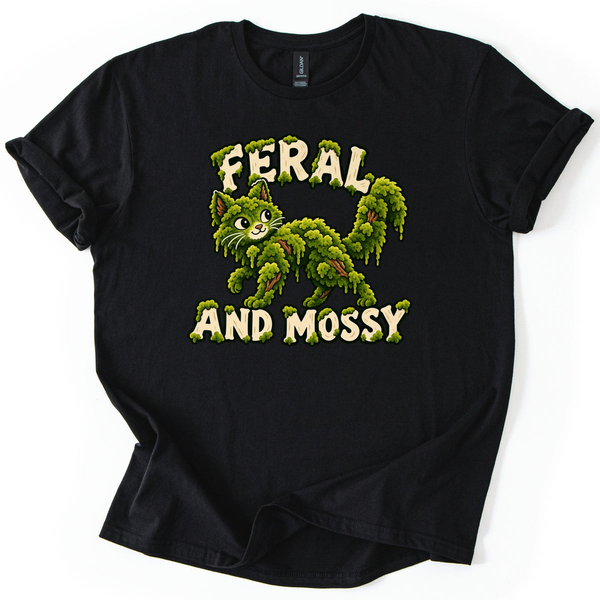 Feral and Mossy Comfort Colors T-shirt in Black, laid flat. A smiling cartoon cat made of dripping green moss with brown twigs showing through, framed by moss-covered cream letters reading Feral and Mossy. The shirt lies on a white surface with cuffed sleeves.