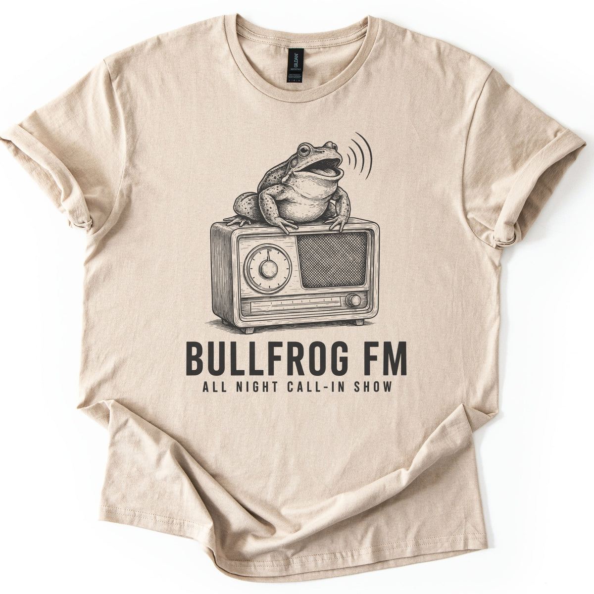 Bullfrog FM Comfort Colors T-shirt in Sand, laid flat. A black ink sketch of a croaking bullfrog on a vintage radio, sound waves at its mouth, above the words BULLFROG FM and ALL NIGHT CALL-IN SHOW. Sleeves are cuffed on a white background.