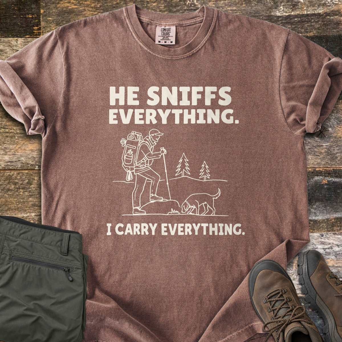 He Sniffs Everything. I Carry Everything Comfort Colors T-shirt in Espresso, laid flat. Cream line art of a hiker with a large backpack and trekking pole beside a dog sniffing a rock, between the words He Sniffs Everything. and I Carry Everything. Olive pants and brown hiking boots sit on weathered wood.