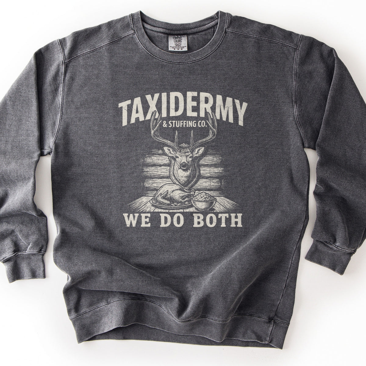 Taxidermy & Stuffing Co. We Do Both Comfort Colors crewneck sweatshirt in Pepper, laid flat. A faded off-white print reads TAXIDERMY & STUFFING CO. over a sketched deer head mounted on a log wall, a roast turkey and a bowl of stuffing on a table, then WE DO BOTH. It lies on a white surface.