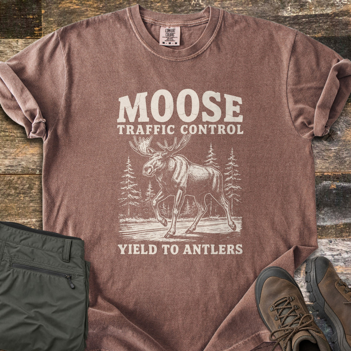 Moose Traffic Control Comfort Colors T-shirt in Espresso, laid flat. An off-white sketch of a moose walking across a road between pine trees, with MOOSE TRAFFIC CONTROL above and YIELD TO ANTLERS below. Sleeves rolled, on weathered wood with olive pants and brown hiking boots.