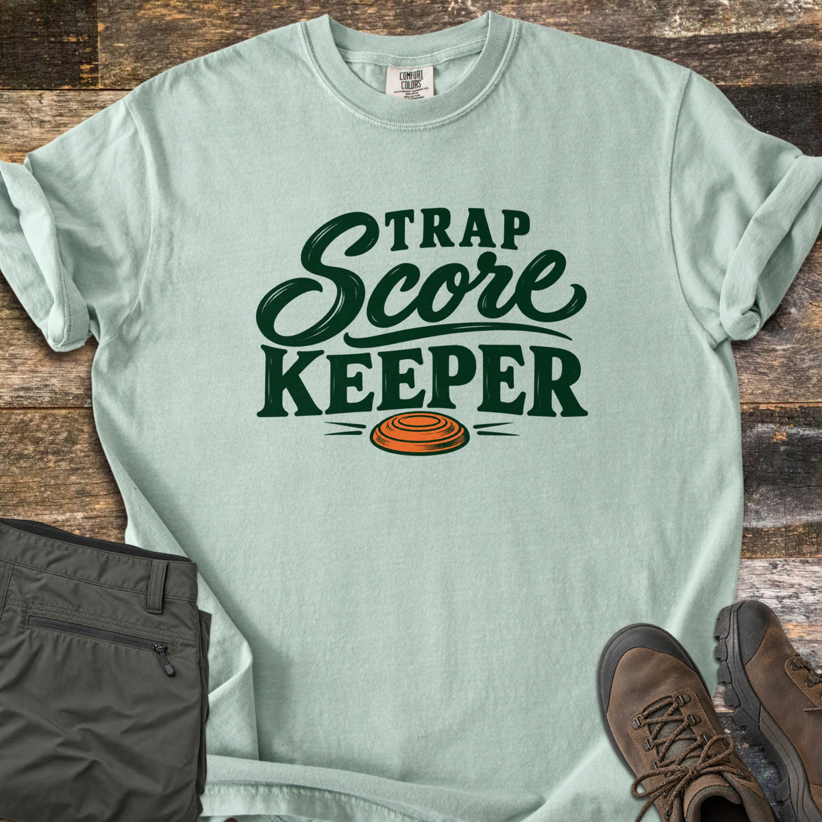 Trap Score Keeper Comfort Colors T-shirt in Bay, laid flat. Dark green lettering reads Trap Score Keeper above an orange clay target. Sleeves are cuffed on weathered wood, with folded olive cargo pants and brown hiking boots beside it.