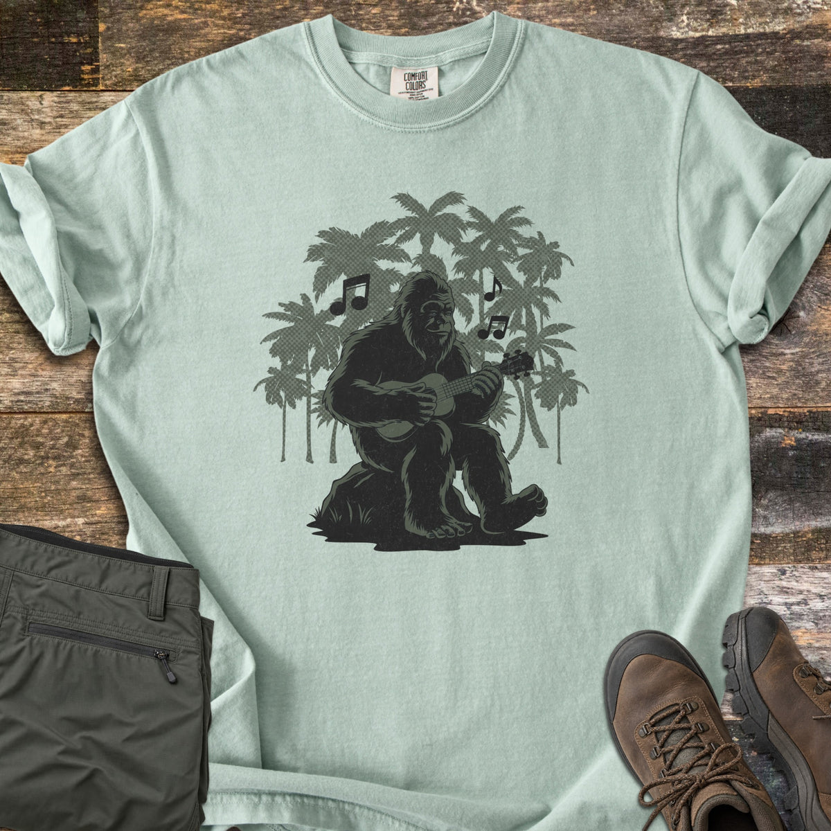 Bigfoot Ukulele Comfort Colors T-shirt in Bay, laid flat. A Bigfoot sits on a rock playing a ukulele, with music notes and green palm tree silhouettes behind him. Olive hiking pants and brown hiking boots lie beside it on weathered wood planks.
