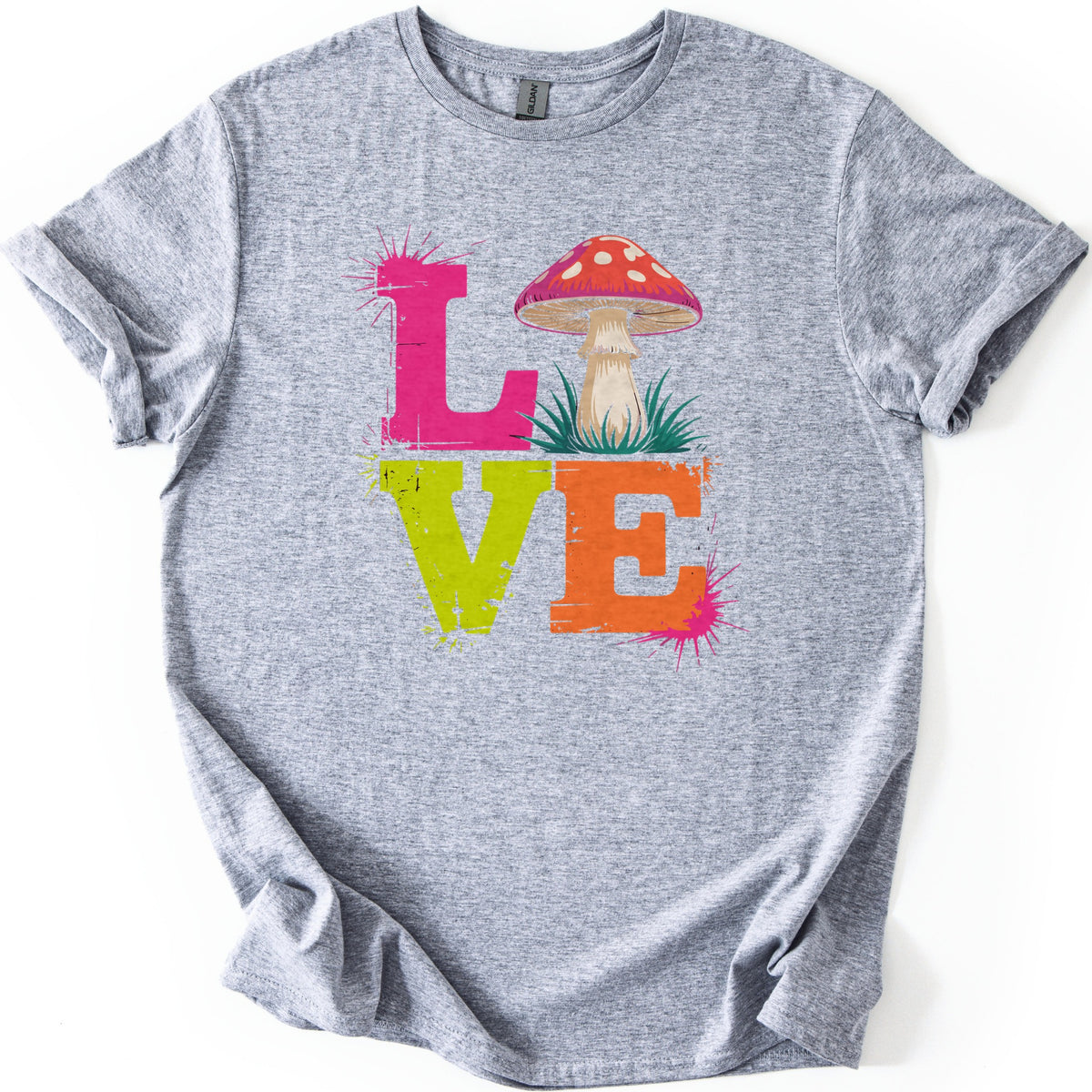 Mushroom Love Comfort Colors T-shirt in Sport Grey, laid flat. The word LOVE in distressed block letters, with a pink L, a red white-spotted mushroom in grass for the O, a lime green V and an orange E, plus pink paint splatters. The shirt lies on white with cuffed sleeves.