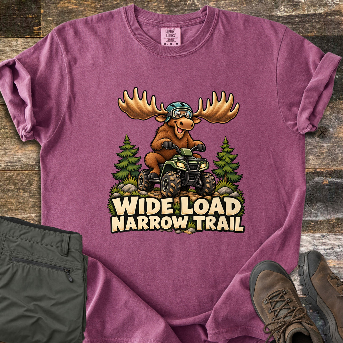 Wide Load Narrow Trail Comfort Colors T-shirt in Berry, laid flat. A grinning cartoon moose in a helmet and goggles drives a green ATV over a rocky trail between pine trees above the words Wide Load Narrow Trail. Olive pants and brown hiking boots sit on weathered wood boards.