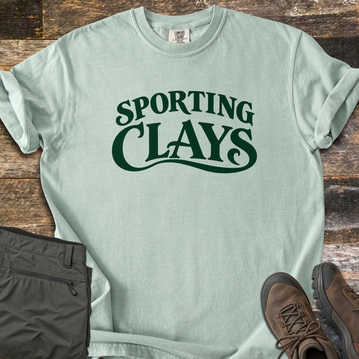 Sporting Clays Comfort Colors T-shirt in Bay, laid flat. Dark green retro lettering reads Sporting Clays, with a swash curving under the word Clays. Sleeves are rolled on a rustic wood floor beside folded olive hiking pants and brown leather hiking boots.